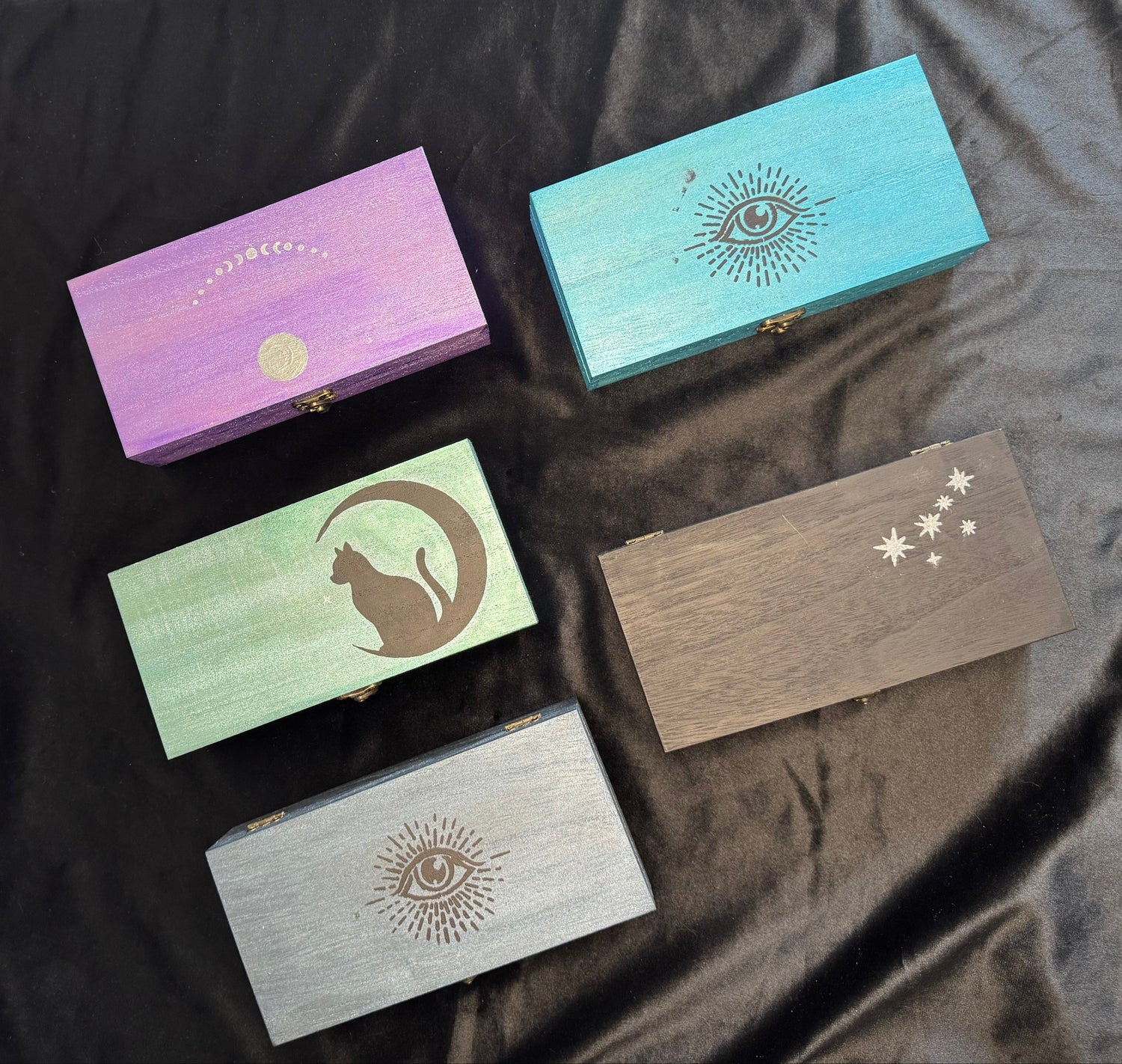 Five rectangular wooden boxes with decorative engravings and lids, displayed on a black satin fabric. The boxes are in different colors: gray, mint green, lavender, brown, and turquoise blue.