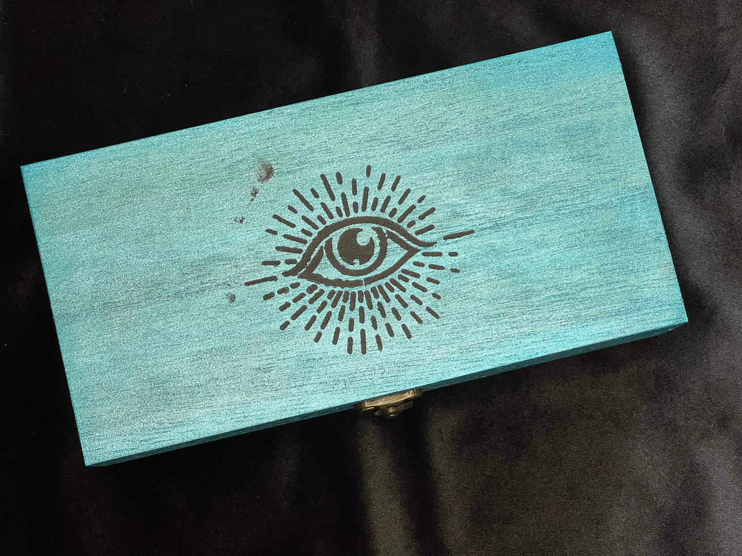 A rectangular light blue wooden box with a black painted eye design on the lid.