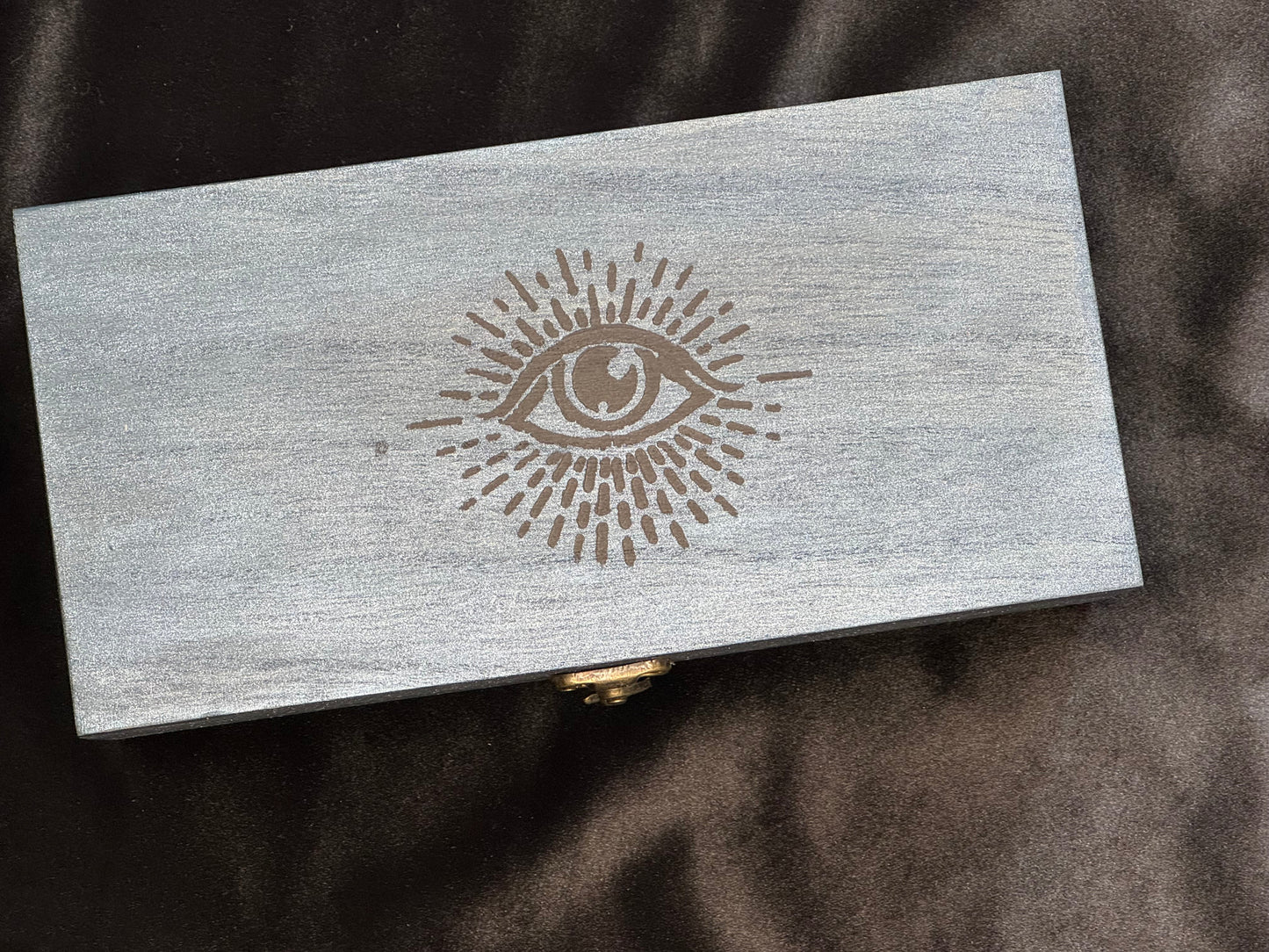 A rectangular metallic navy wooden box with a black painted eye surrounded by rays on the lid.