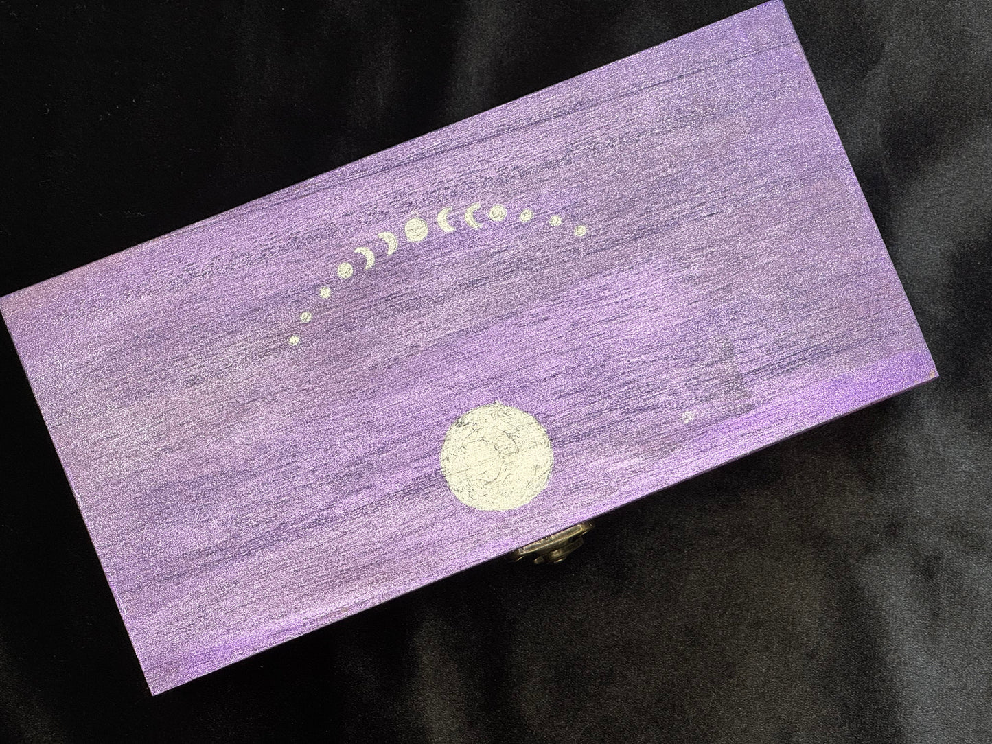 A rectangular purple box with a distressed wood-grain texture, decorated with a silver moon phase design and a small brass clasp.