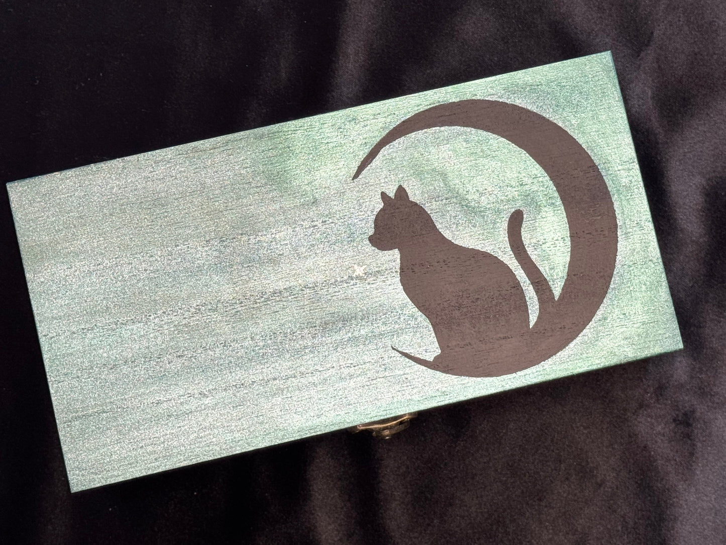 A rectangular wooden box with a teal metallaic finish, featuring a black silhouette of a cat sitting inside a crescent moon on the front.